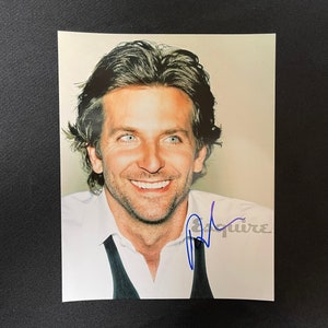 May include: A colour photograph of a man with short brown hair smiling. He is wearing a white shirt with a black bow tie. The man's signature is in blue ink below the image. The signature reads "Bradley Cooper Esquire".