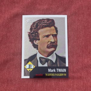 Könnte beinhalten: Eine silberne und weiße Sammelkarte mit einem Porträt von Mark Twain, einem Romancier. Die Karte hat ein rotes und gelbes Logo mit einer Feder und einem offenen Buch. Der Text auf der Karte lautet "Mark Twain, Die Abenteuer des Huckleberry Finn, Romancier."