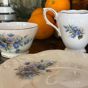 May include: A white porcelain tea set with a floral pattern of blue flowers and gold edging. The set includes a creamer, a sugar bowl, and a serving plate. The floral design is complemented by pink and green accents.