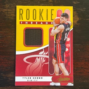 Op de afbeelding: Een basketbal trading card met Tyler Herro van de Miami Heat. De kaart is geel en rood met een zwarte jersey patch. Op de kaart staat de tekst "Rookie Threads" en "Tyler Herro Miami Heat" gedrukt.