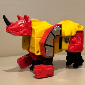 May include: A red and yellow toy robot in rhinoceros form. The robot has a black horn and black legs. The robot has a silver panel on its side.