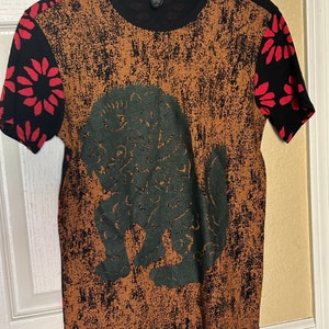 May include: A black and brown short-sleeved t-shirt with a green graphic print of a mythical creature. The sleeves have a red floral pattern on a black background.