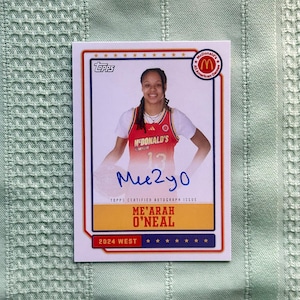Puede incluir: Una tarjeta comercial Topps con Me'Arah O'Neal con una camiseta roja y blanca de McDonald's. La tarjeta tiene un borde blanco con detalles en rojo y azul, el logotipo de McDonald's All American Games y su autógrafo. La tarjeta dice "2024 West".