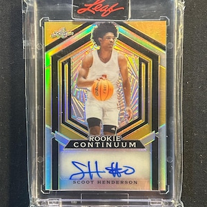 May include: A basketball trading card in a protective sleeve. The card shows a player with a basketball, with the text "ROOKIE CONTINUUM" and a signature. The card has a gold and iridescent design with the "Leaf" logo.
