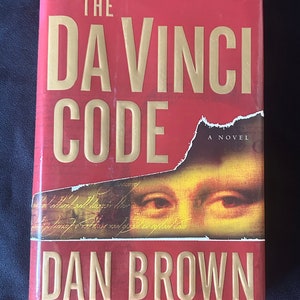 Könnte beinhalten: Der Da Vinci Code von Dan Brown, ein Roman. Das Cover zeigt einen roten Hintergrund mit goldenen Buchstaben und eine Nahaufnahme der Augen der Mona Lisa.