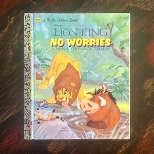 May include: A Little Golden Book titled "The Lion King No Worries: A New Story About Simba." The cover features Simba, Pumbaa, and Timon in a colorful illustration. The book has a gold spine and is set against a wooden background.