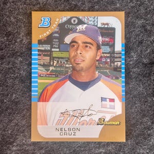 May include: A baseball card featuring Nelson Cruz in a baseball uniform. The card has a gold border with blue stripes and the Bowman logo. The text "FIRST YEAR" is visible, along with Cruz's signature and name.