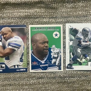 May include: Three trading cards featuring Emmitt Smith, a former NFL running back for the Dallas Cowboys. The cards are from different sets and feature different photos of Smith. The first card is from the XL set and features a photo of Smith in a blue and white uniform. The second card is from the Fleer Tradition set and features a photo of Smith in a blue and white uniform with a green background. The third card is from the SP Authentic set and features a photo of Smith in a blue and white uniform running with the ball.