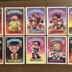 Może przedstawiać: Zestaw dziewięciu kart kolekcjonerskich z postaciami z kreskówek z serii Garbage Pail Kids. Karty są białe z kolorową ramką i przedstawiają różne postacie o imionach takich jak Bad Breath Seth, Foul Phil, Hairy Gary, Hairy Carrie, Beth Death, Spacey Stacy, Russell Muscle, Marty Gras, Outer Space Chase i Luke Warm.