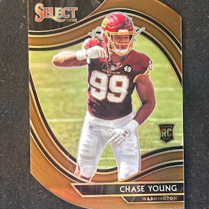 May include: A gold and black football trading card featuring a photo of Chase Young, a Washington Football Team player, wearing a burgundy and gold uniform with the number 99. The card is labelled "SELECT 2020" and "Rookie RC Card".