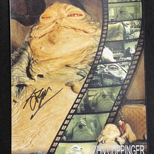 May include: A black and white photo of a Jabba the Hutt puppet from the Star Wars movie. The photo is signed by John Coppinger, a special effects artist who worked on the film. The photo also includes a film strip with images of the puppet being constructed.