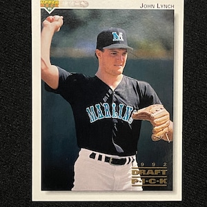 Puede incluir: Una tarjeta de béisbol con un jugador de béisbol que lleva un uniforme negro y verde azulado con la palabra "Marlins" en la parte delantera. El jugador lleva una gorra de béisbol verde azulado con una "M" blanca en la parte delantera. La tarjeta es de la serie Upper Deck de 1992 y presenta el texto "John Lynch" y "Draft Pick".