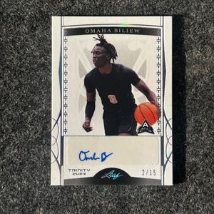 May include: A white and silver basketball trading card featuring Omaha Biliew, a basketball player in a black jersey with a gold number 5. The card has a silver border and a blue signature. The card is numbered 2/15.