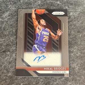 May include: A basketball trading card featuring Mikal Bridges of the Phoenix Suns. The card has a silver, black, and red design with a white autograph patch. The card features a photo of Bridges in action, and his signature is on the autograph patch. The card is from the Panini Prizm series.
