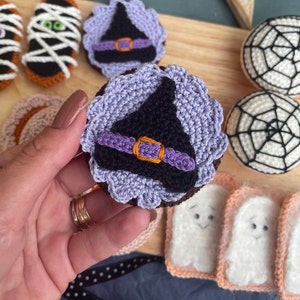 May include: A close-up of a crocheted witch hat cookie. The hat is purple and black with an orange buckle. The cookie is surrounded by other crocheted Halloween treats, including ghosts, spiderwebs, and mummies.