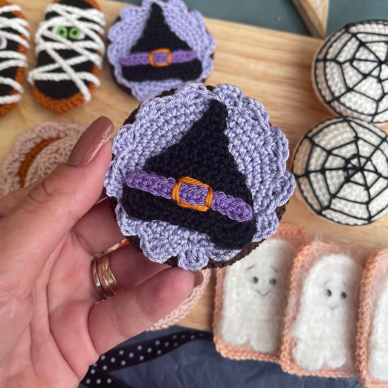 May include: A close-up of a crocheted witch hat cookie. The hat is purple and black with an orange buckle. The cookie is surrounded by other crocheted Halloween treats, including ghosts, spiderwebs, and mummies.