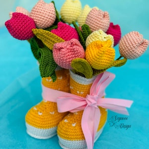 May include: A crocheted bouquet of pink, yellow, and red tulips in a yellow crocheted boot with a pink ribbon. The boots are on a blue surface. The text "Saymoo Design" is visible in the lower right corner of the image.