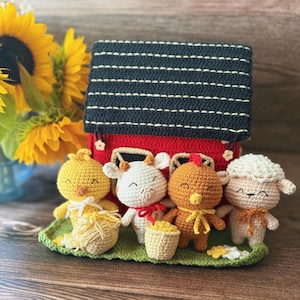 May include: A crochet barn set featuring a red barn with a dark roof and four small stuffed animals: a yellow chick, a white cow, a brown chicken, and a cream sheep. The figures stand on a green base, each with a small accessory like a basket or bow.