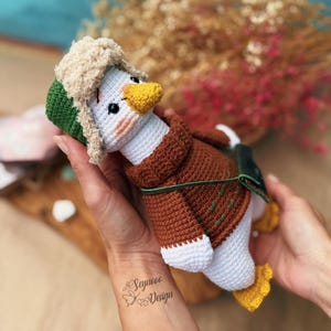 May include: A handmade crochet duck wearing a brown jacket, green and beige hat, and yellow feet. The duck has a white body, a yellow beak, and black eyes. The duck is held in two hands.