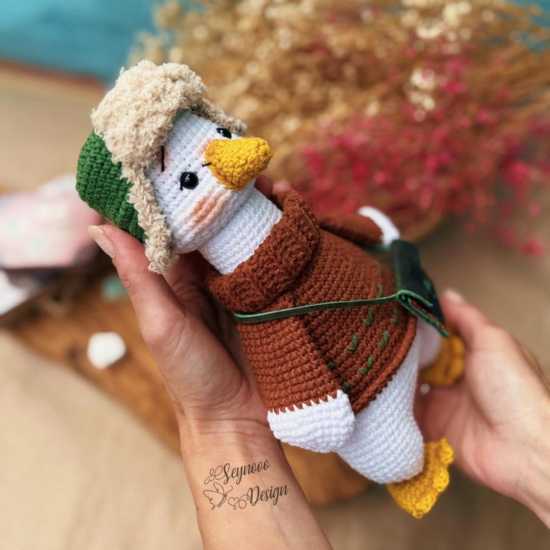 May include: A handmade crochet duck wearing a brown jacket, green and beige hat, and yellow feet. The duck has a white body, a yellow beak, and black eyes. The duck is held in two hands.