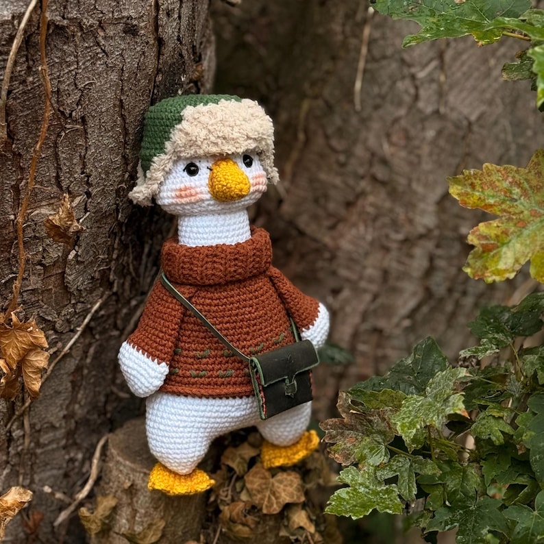 May include: A handmade crochet goose doll wearing a brown sweater, green hat with ear flaps, and a small green bag. The goose has a yellow beak and feet, and is standing on a tree stump. The doll is a whimsical, handcrafted item.