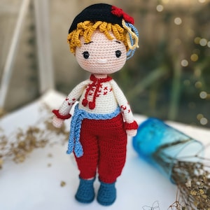 May include: A crocheted doll wearing a black hat with a red flower, a white shirt with red and blue embroidery, red trousers, and blue shoes. The doll has blonde hair and a sweet expression.