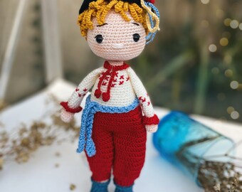 Puede incluir: Una muñeca de crochet que lleva un sombrero negro con una flor roja, una camisa blanca con bordados rojos y azules, pantalones rojos y zapatos azules. La muñeca tiene el pelo rubio y una expresión dulce.