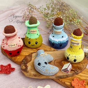 Könnte beinhalten: Fünf gehäkelte Amigurumi-Flaschen in verschiedenen Farben und mit Gesichtern. Jede Flasche hat ein Etikett mit einem anderen Wort: Liebe, Weisheit, Macht, Mut und Traum. Die Flaschen stehen auf einer Holzoberfläche.