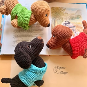 May include: Crocheted dachshund dog toys in various colors, including brown, black, and tan, each wearing a sweater. The dogs are positioned around an open book. The image includes the text "Seynooo Design".