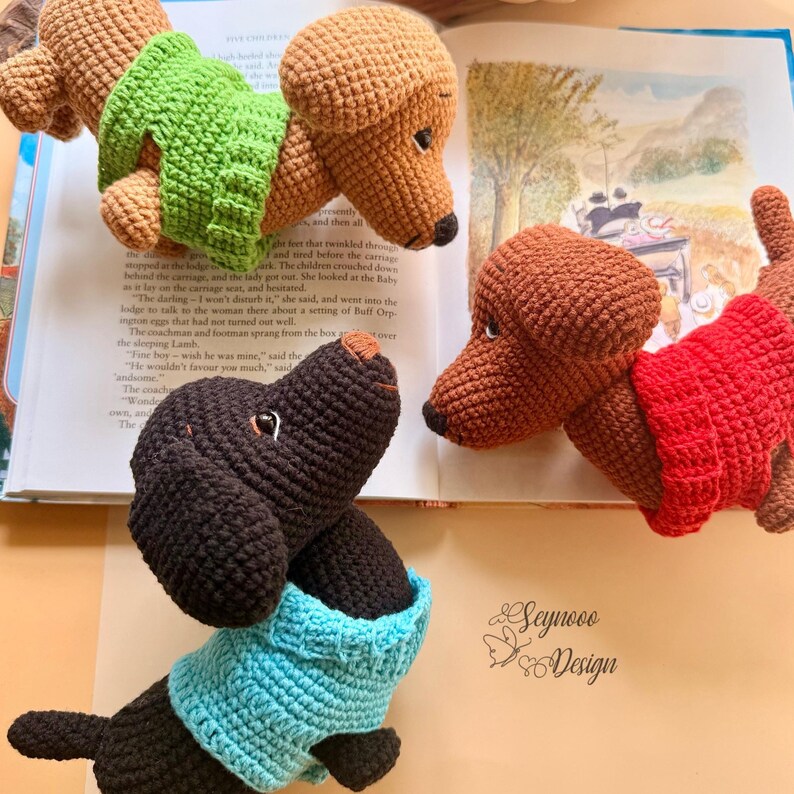 May include: Crocheted dachshund dog toys in various colors, including brown, black, and tan, each wearing a sweater. The dogs are positioned around an open book. The image includes the text "Seynooo Design".