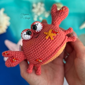 Pode incluir: Um caranguejo vermelho de crochê com uma estrela do mar amarela nas costas. O caranguejo tem olhos pretos grandes e é decorado com contas laranja e vermelhas.