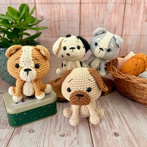 May include: Four handmade crochet dog figurines in various colors: tan and white, black and white, gray and white, and tan and brown. The dogs are sitting on a wooden surface with a green tin and a basket of yarn in the background. The dogs are approximately 4 inches tall.