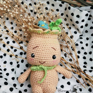 Può includere: Una bambola amigurumi all'uncinetto a forma di pianta con una sciarpa verde e un piccolo nido di uccelli sulla testa. La bambola è seduta su un tessuto bianco con pois neri.