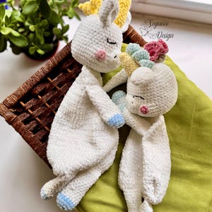 Puede incluir: Dos muñecas de unicornio blancas hechas a mano con ganchillo, con los ojos cerrados, mejillas rosas y crines coloridas. Un unicornio tiene una pezuña azul, y el otro tiene una crin amarilla, azul y rosa. Las muñecas están sobre una superficie verde.