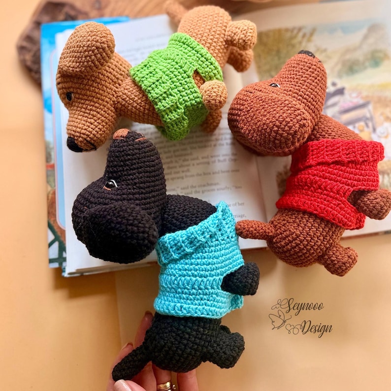 May include: Crocheted dachshund dog toys in brown and black, each wearing a different colored sweater: green, red, and blue. The toys are handmade and sit on top of an open book. The image includes the text "Seyneco Design".
