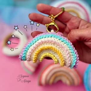 Peut inclure: Un porte-clés en crochet en forme d'arc-en-ciel avec un anneau de clé en métal doré. Le porte-clés est composé de cinq bandes de couleurs différentes. Le porte-clés est tenu dans une main sur un fond rose.