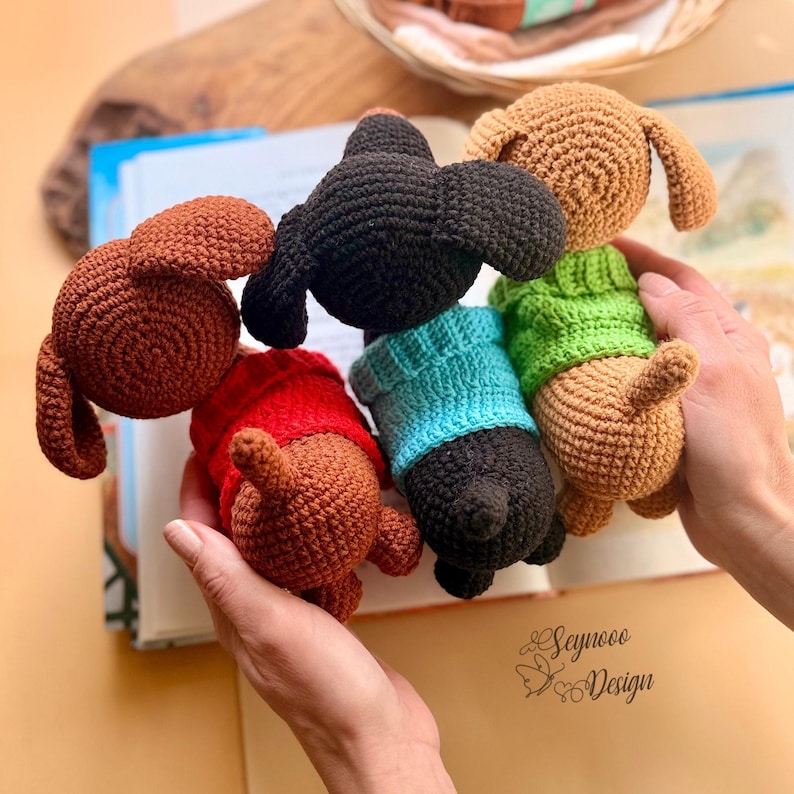 May include: A collection of five crocheted dachshund dog toys in various colors. The dogs are brown, black, and tan, with some wearing sweaters in red, blue, and green. The toys are held in a person's hands, with a book in the background.