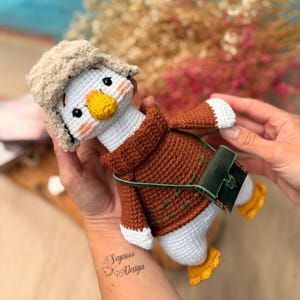 May include: A handmade crochet goose doll wearing a brown sweater, a beige hat, and a green leather bag. The goose has a yellow beak and feet, with pink cheeks. The doll is held in a person's hands.
