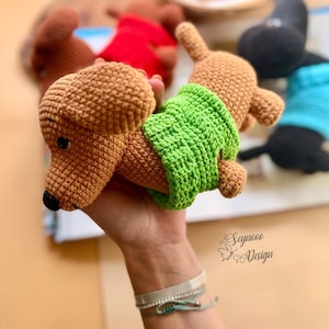 May include: A crocheted dachshund dog toy in shades of brown, with a green crocheted belly band. The dog has a black nose and small ears. Other similar toys in red and blue are visible in the background.