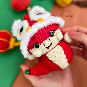 Puede incluir: Un disfraz de danza de león rojo y blanco de ganchillo con una cara sonriente y ojos grandes. El disfraz está hecho de hilo y tiene una melena blanca esponjosa. El león sostiene una pequeña bola roja en su pata.