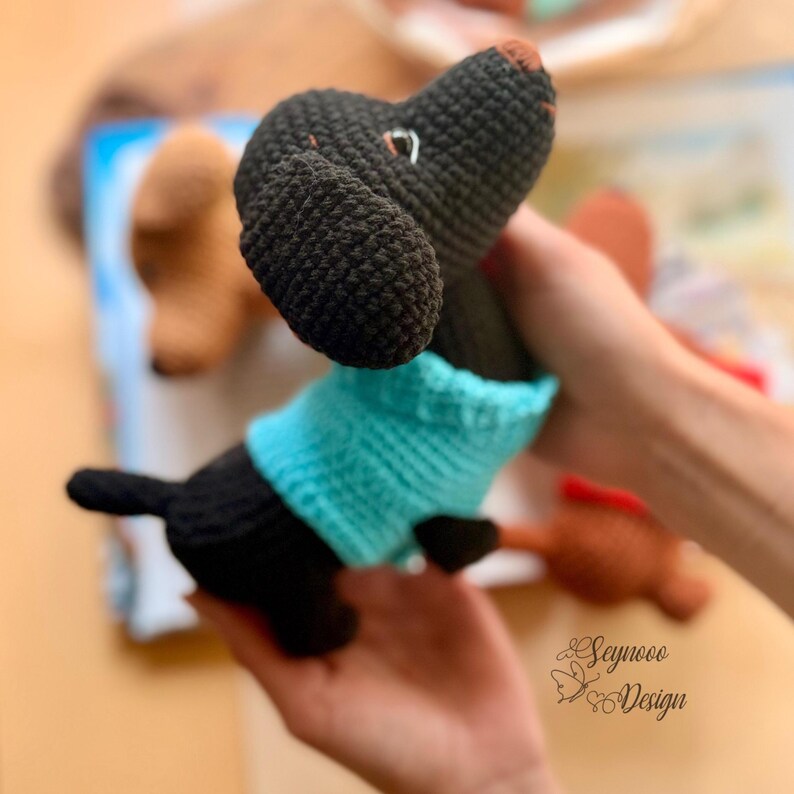 May include: A handmade crochet dachshund dog in black and teal. The dog has a teal sweater and a brown nose. The dog is being held in someone's hand. The dog is a cute and cuddly toy.