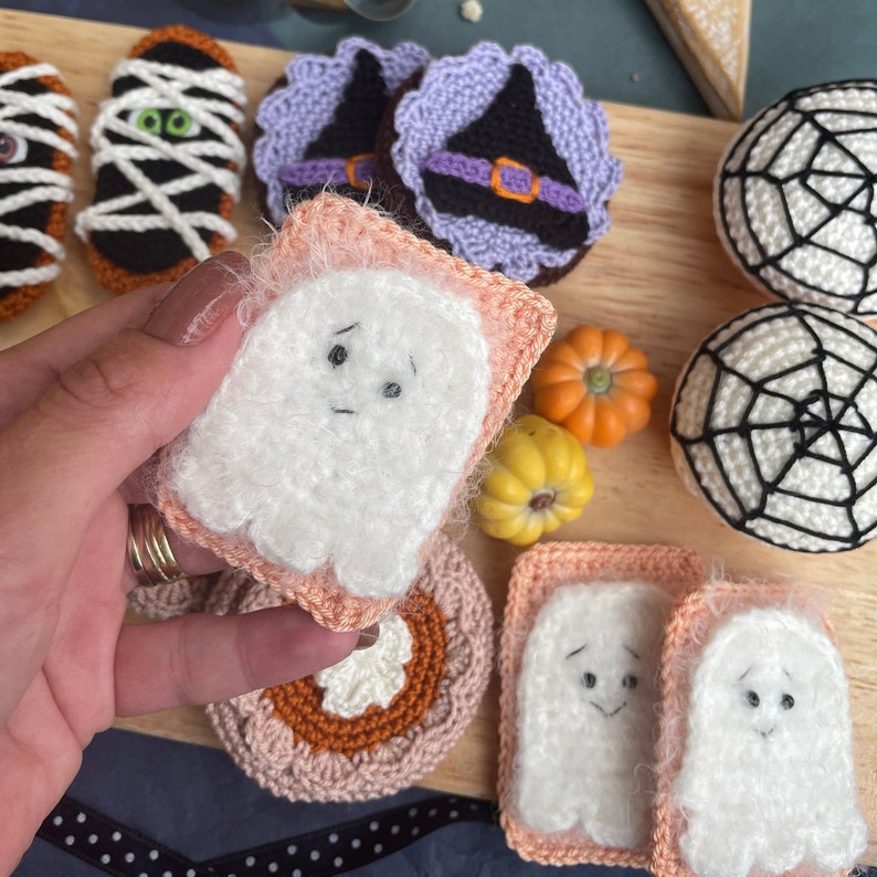May include: A collection of crocheted Halloween decorations, including a ghost, a witch's hat, a spiderweb, and a pumpkin. The decorations are made with white, purple, black, and orange yarn.