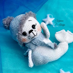 Puede incluir: Una foca de crochet blanco con un gorro y una bufanda de punto azul. La foca tiene un pompón blanco esponjoso en su gorro y está tumbada sobre una superficie azul.