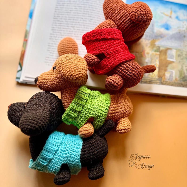 May include: A collection of crocheted dachshund dog figurines in various colors, including brown, black, and tan. Some dogs wear tiny sweaters in red, green, and blue. The image includes a partially visible open book.