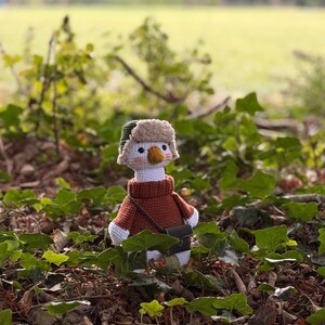 May include: A handmade crochet goose doll wearing a brown sweater, a green and tan hat, and a small black bag. The goose has a white body, a yellow beak, and is surrounded by green foliage.