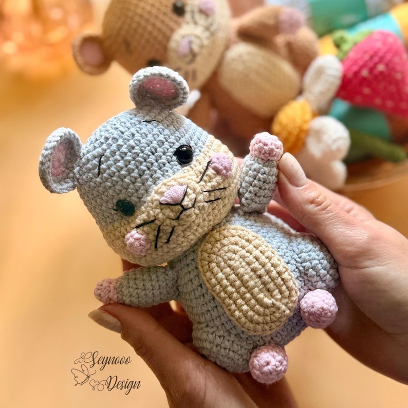 May include: A handmade crochet mouse toy in shades of gray, beige, and pink. The mouse has small black eyes, pink ears, and a pink nose. The toy is held in a person's hands. The text "Seynooo Design" is visible.