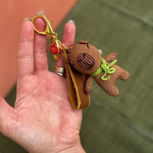 May include: A brown crocheted teddy bear keychain with a green scarf and a brown leather strap. The keychain is attached to a gold metal ring.