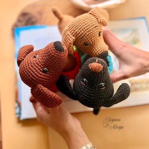 May include: Three handmade crochet dog toys in brown, tan, and black. The toys have black button eyes and are held over an open book. The toys are a charming addition to a child's toy collection.