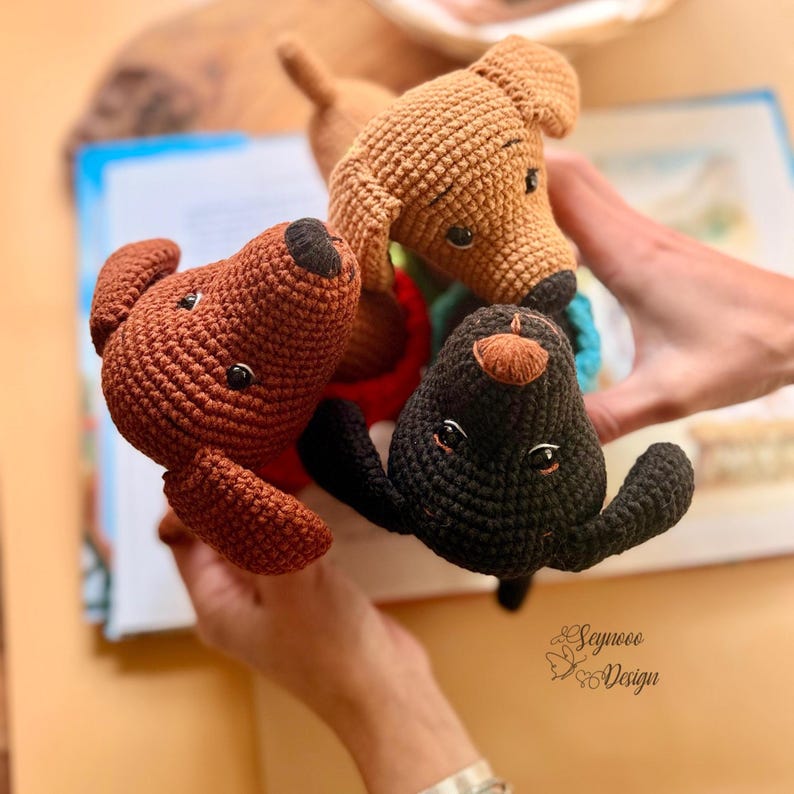 May include: Three handmade crochet dog toys in brown, tan, and black. The toys have black button eyes and are held over an open book. The toys are a charming addition to a child's toy collection.