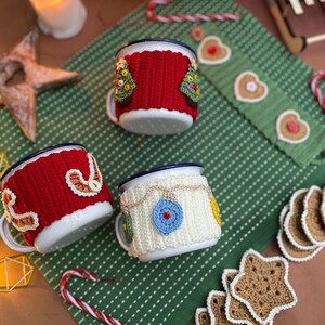 May include: Three white enamel mugs with blue rims, each adorned with a unique, colorful, crocheted cozy. One mug features a red cozy with a Christmas tree design, another with a white cozy and ornament design, and the third with a red cozy and button design.